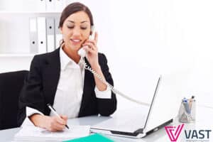 Happy Executive Assistant on the phone in front of her desk dressed in a white blouse and black blazer.