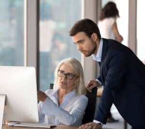 A young white executive in a 3-piece suit leaning over a desk looking at a computer screen with a middle-aged female executive assistant explaining what is on the computer screen.