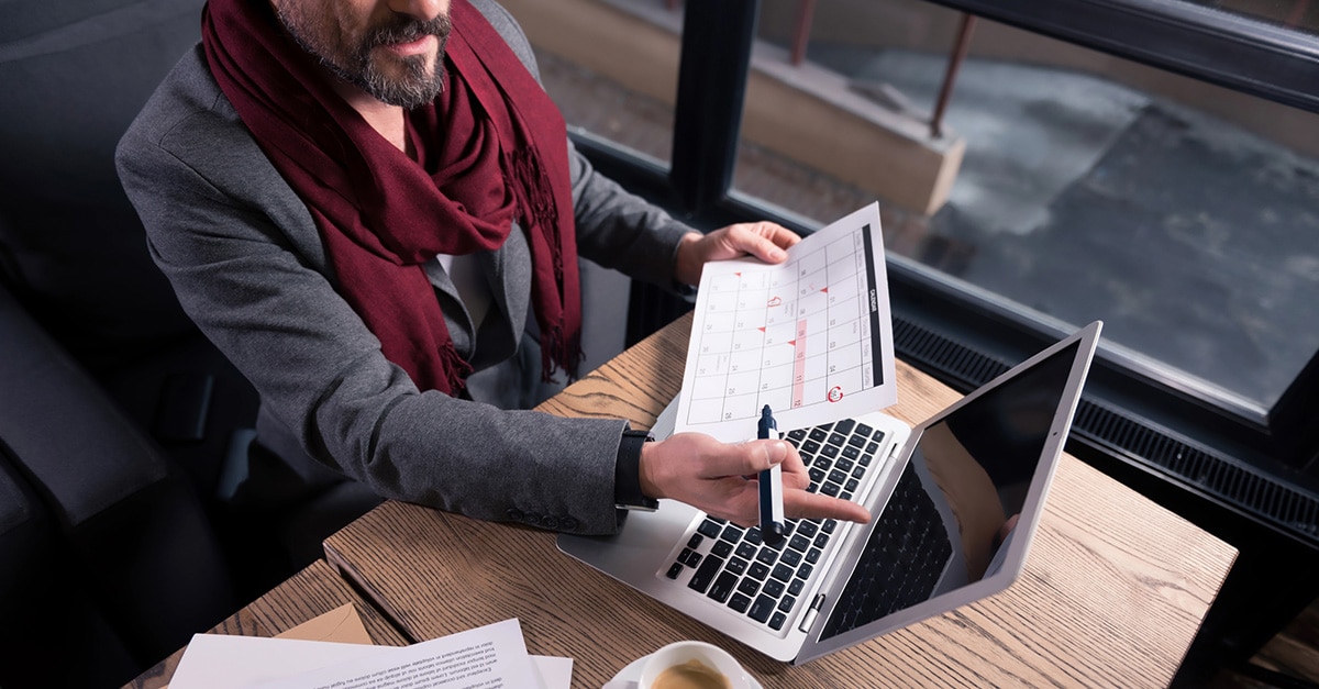 The image shows a person in business attire holding a calendar and pointing to a laptop screen with a pen, suggesting planning, scheduling, or organizing activities. The setting appears to be a professional or casual office environment with natural light from a nearby window. A cup of coffee and papers are on the desk, reinforcing a work-focused context.