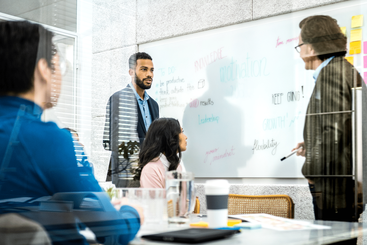 A group of individuals gathered in a meeting room, focused on a whiteboard while discussing ideas and strategies.