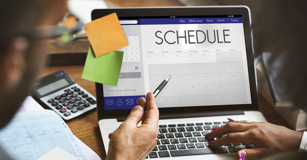 This image shows a laptop displaying a digital calendar with the word "SCHEDULE" prominently at the top. A person is pointing at the screen with a pen, suggesting active planning or adjustments. Office items like a calculator and sticky notes surround the laptop, reinforcing the context of professional scheduling and organization. The image aligns with the topic of managing last-minute scheduling changes effectively.