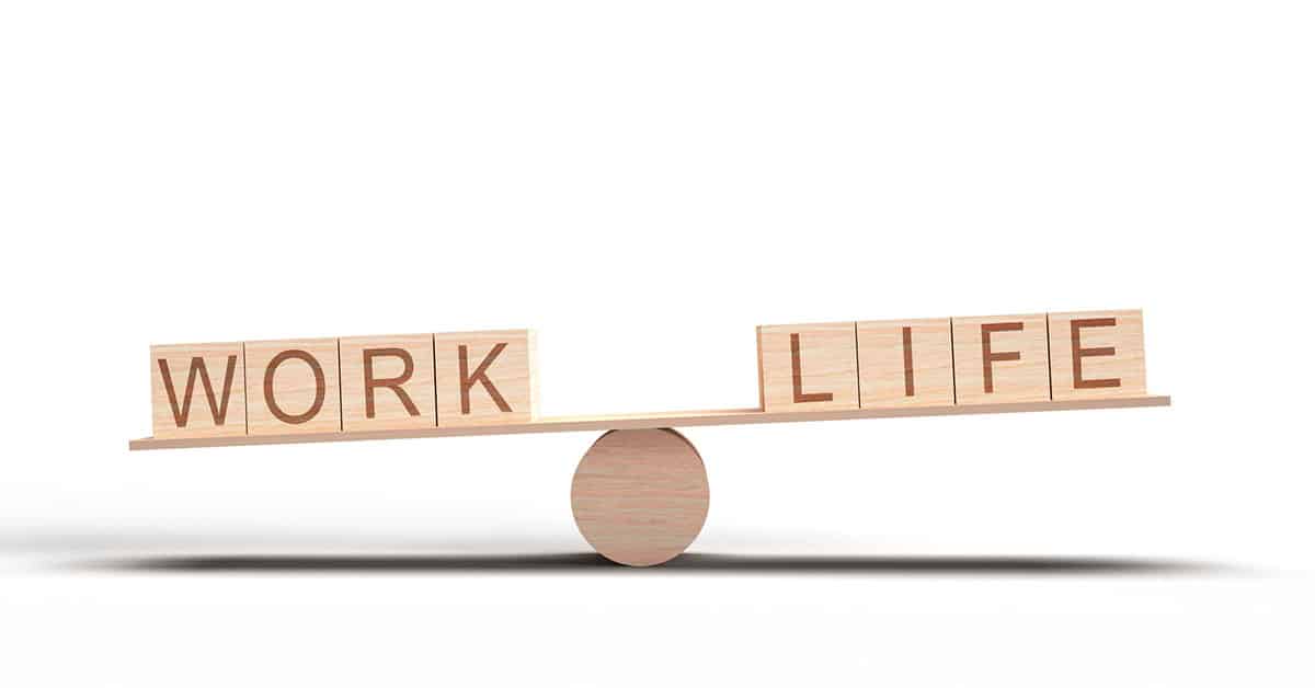 The image depicts a wooden seesaw balanced on a cylindrical pivot, with the words "WORK" on one side and "LIFE" on the other. The seesaw is slightly tilted, indicating an imbalance. This visual metaphor represents the challenge of maintaining a work-life balance, especially relevant for professionals like executive assistants who juggle multiple responsibilities. Let me know if you need an alt text description or any edits for accessibility purposes.