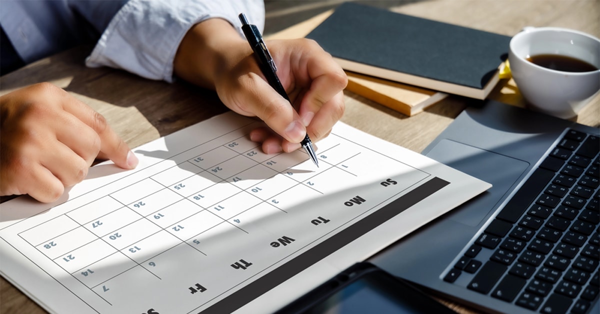 A person reviewing a printed calendar with a pen in hand, pointing at a date. The calendar is placed on a wooden desk alongside a laptop, a tablet with a stylus, a cup of coffee, and notebooks. Sunlight casts shadows across the workspace, indicating a well-lit environment