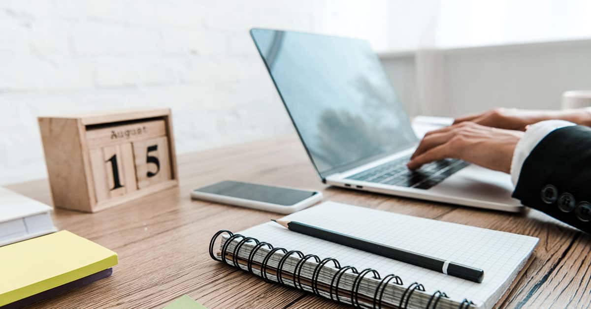 A professional workspace with a wooden desk, featuring a laptop, smartphone, spiral notebook with a pen, and a wooden calendar displaying August 15. A person in a suit is typing on the laptop, suggesting productivity and organization.