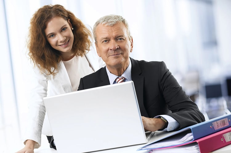 A professional executive in a suit working on a laptop, with a smiling assistant standing beside him in a modern office setting. The image represents efficient collaboration, highlighting the benefits of using executive assistant software like VAST Calendar Management System to streamline scheduling and productivity.